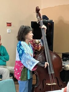 Becky, wearing a colorful top, plays Spirit of Life on her upright bass.